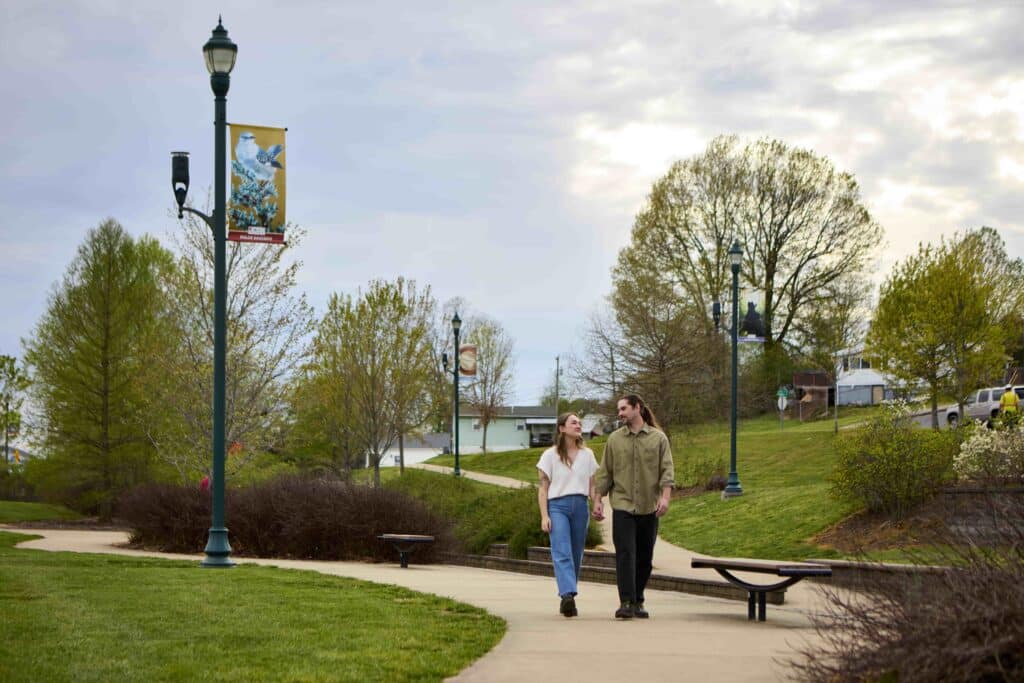 Two people holding hands and walking on a paved park path surrounded by trees and lampposts under a cloudy sky.